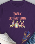 The Easily Distracted By Dogs Series, Volume 2 Womens T-Shirt in purple features the phrase in pink with a Dalmatian, a Golden Retriever, a Pug, a Poodle, Dachshund, French Bulldog, Chihuahua, Husky and more around the letters. Designed by Dog Gang.