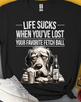 Unisex-T-Shirt mit einem lustig gezeichneten traurigen Hund mit eine Flasche (Flaschentikett: "Chicken Soup with whipped cream, for sad dog days") und dem Text „Life Sucks When You've Lost Your Favorite Fetch Ball“. Design von Dog Gang.