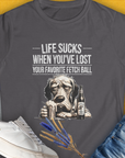 Charcoal t-shirt "Life Sucks When You've Lost Your Favorite Fetch Ball", Volume 5 from the popular series of the same name. Funny drawing of a depressed dog face. Next to the dog is a full glass and a bottle with a label that says "Chicken Soup with Whipped Cream, for Sad Dog Days". Designed by Dog Gang.