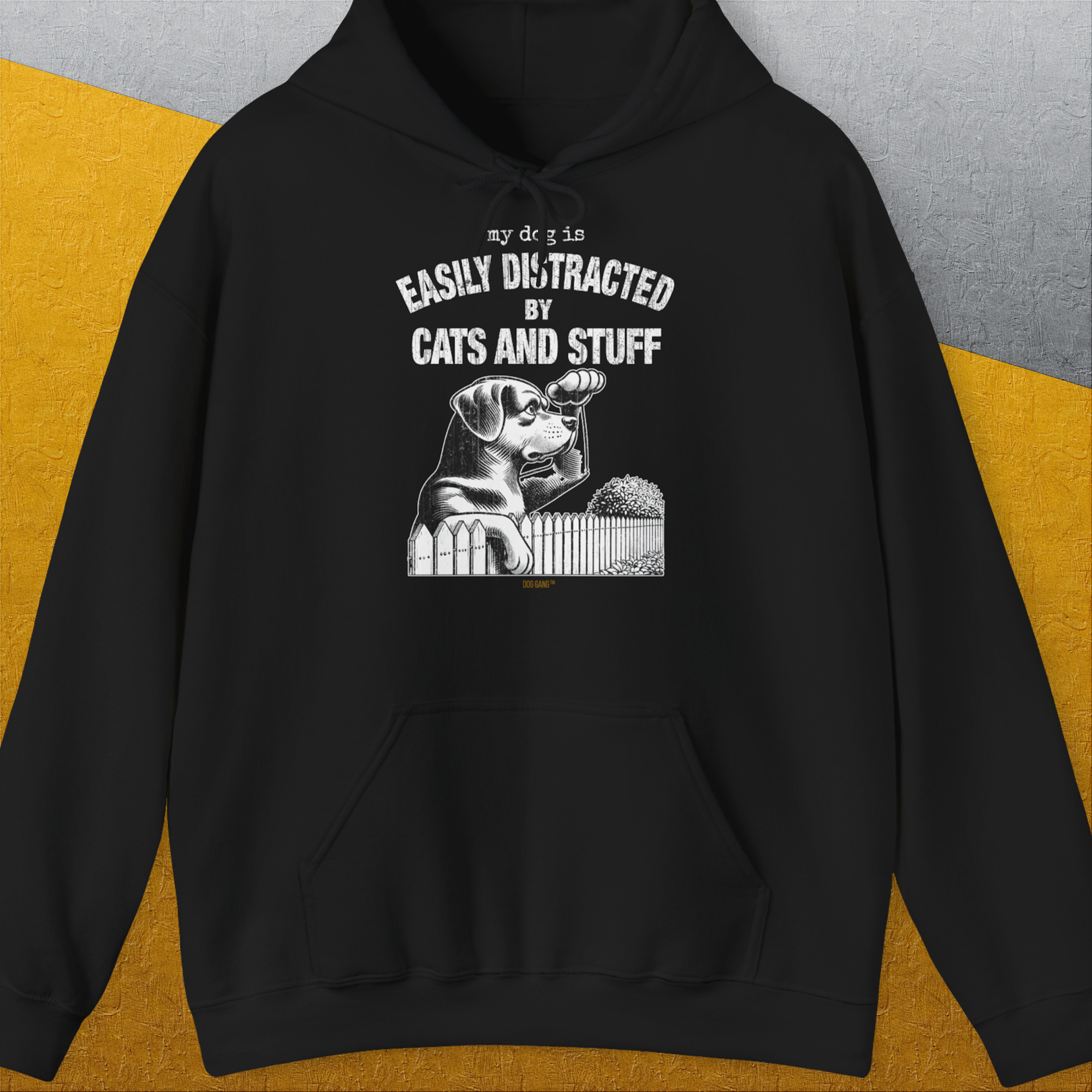 The shirt is a black unisex piece crafted from a cozy cotton-polyester blend. It showcases a charming design of a dog peering curiously at a cat from behind a fence. Bold white letters spell out My dog is easily distracted by cats and stuff, set against textured gray and yellow backgrounds. Shirtdesign by Dog Gang.