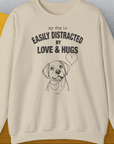 Unisex sand sweatshirt, ethically crafted from cotton, showcases My Dog is Easily Distracted by Love & Hugs text with a delightful dog and heart illustration. Designed by Dog Gang.