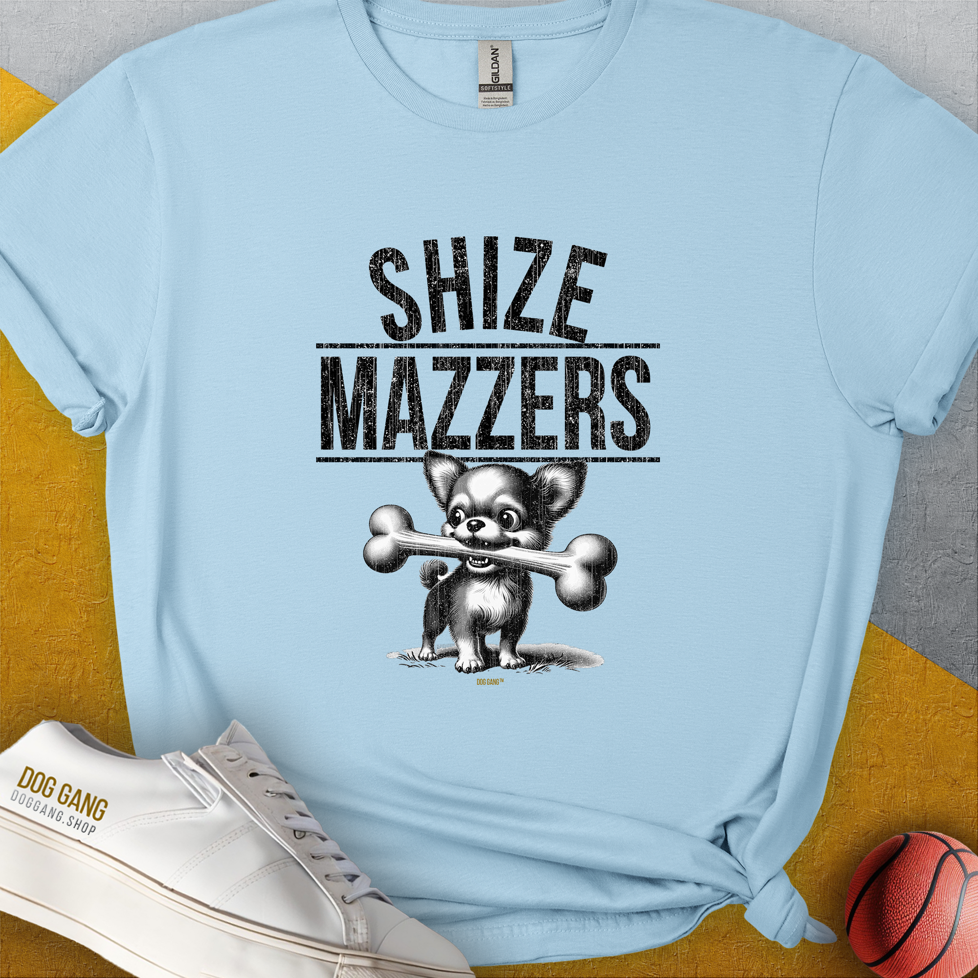 The light blue t-shirt displays a funny cartoon dog holding an oversized bone in its mouth, preventing it from speaking properly. The text reads "SHIZE MAZZERS". Perfect for any dog lover. Designed by Dog Gang.