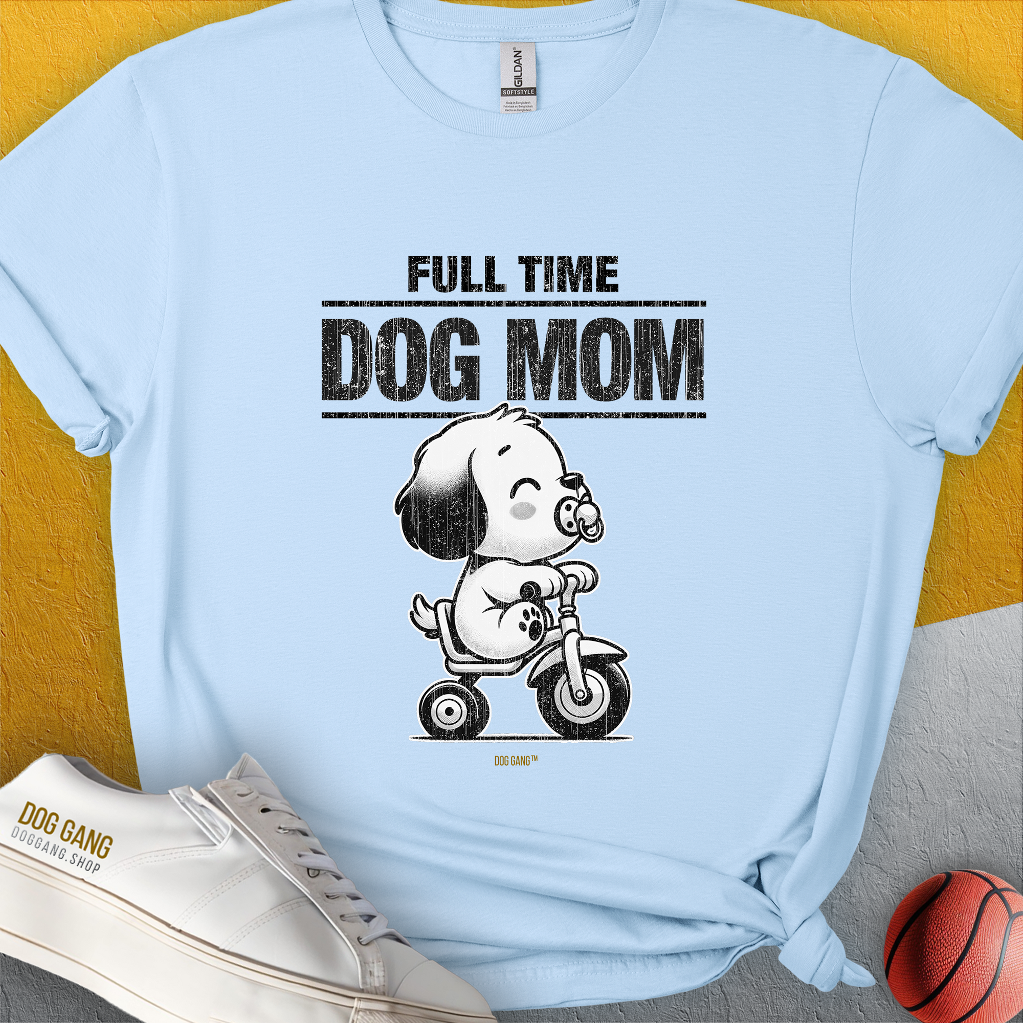 Soft light blue tee with Full Time Dog Mom and a cartoon puppy on a tricycle, playfully surrounded by a ball and shoes—ideal for any dog person. Designed by Dog Gang.