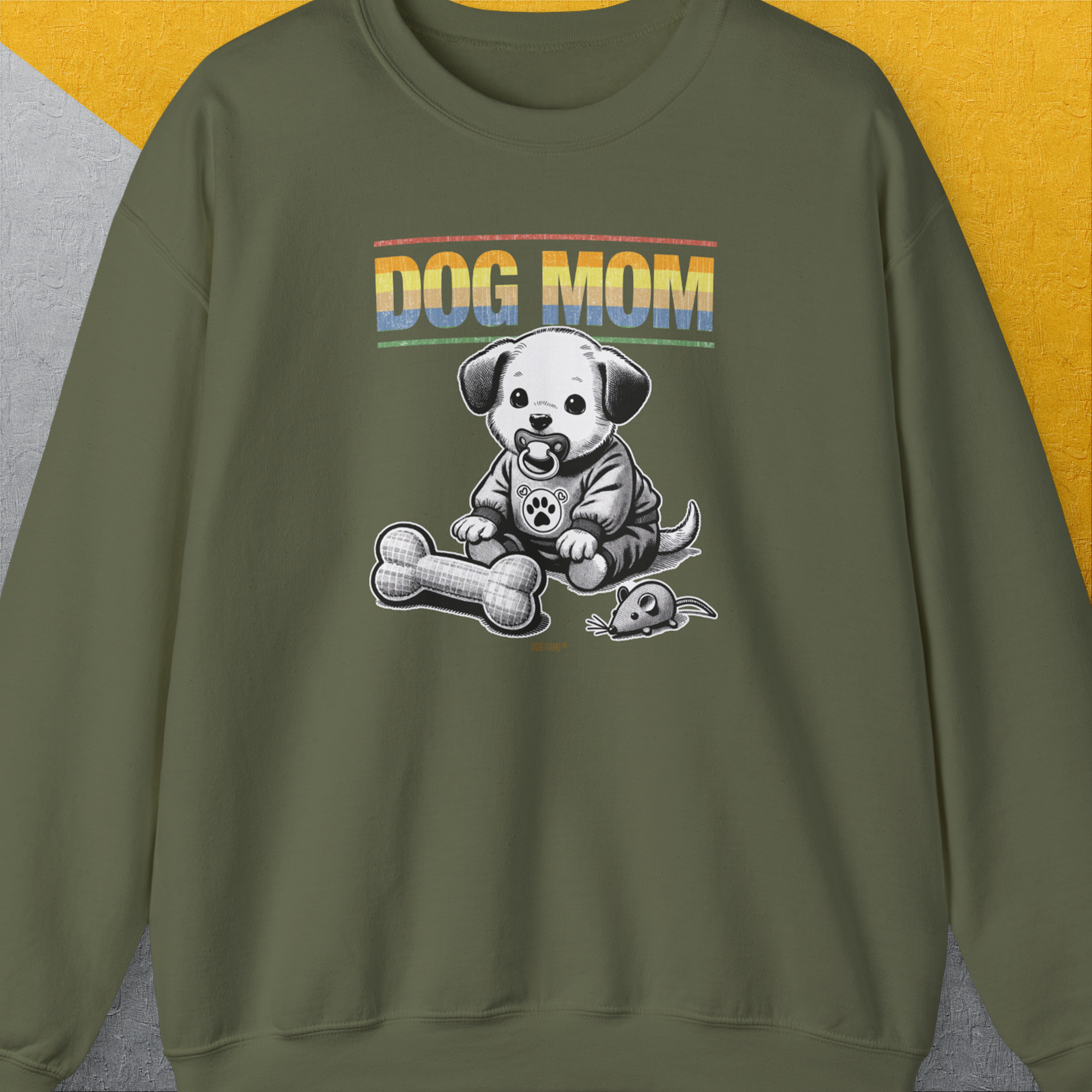 The sweatshirt features a cartoon puppy with a fabric bone and fabric mouse, with Dog Mom text in a sunset gradient. Perfect for any dog person. Designed by Dog Gang.