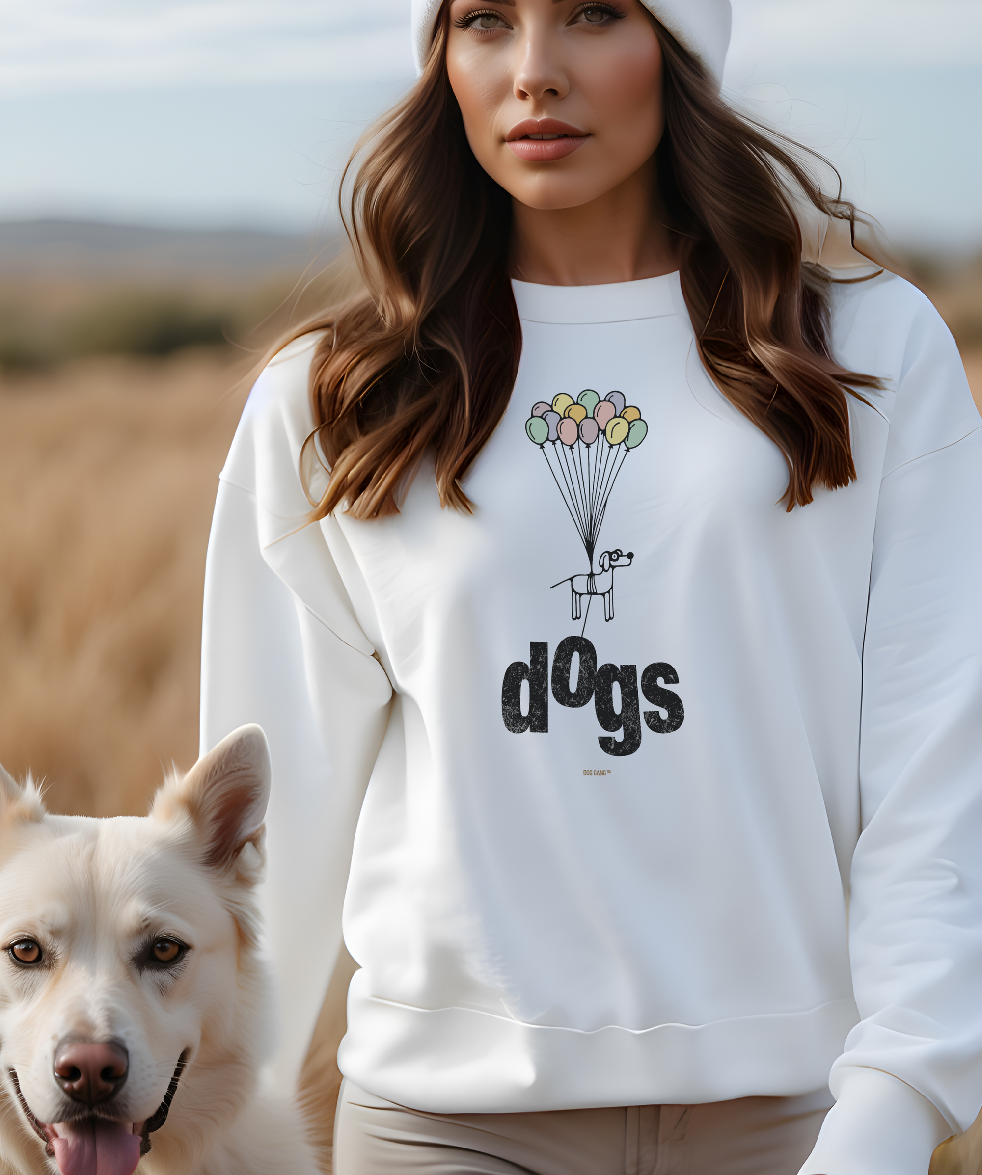 A woman wearing a white sweatshirt from the Designer Label "DOG GANG" with a dogs graphic featuring a funny cartoon dog and balloons stands outdoors. She is next to a light-colored dog. She also wears a white beanie and the background is a field of grass.