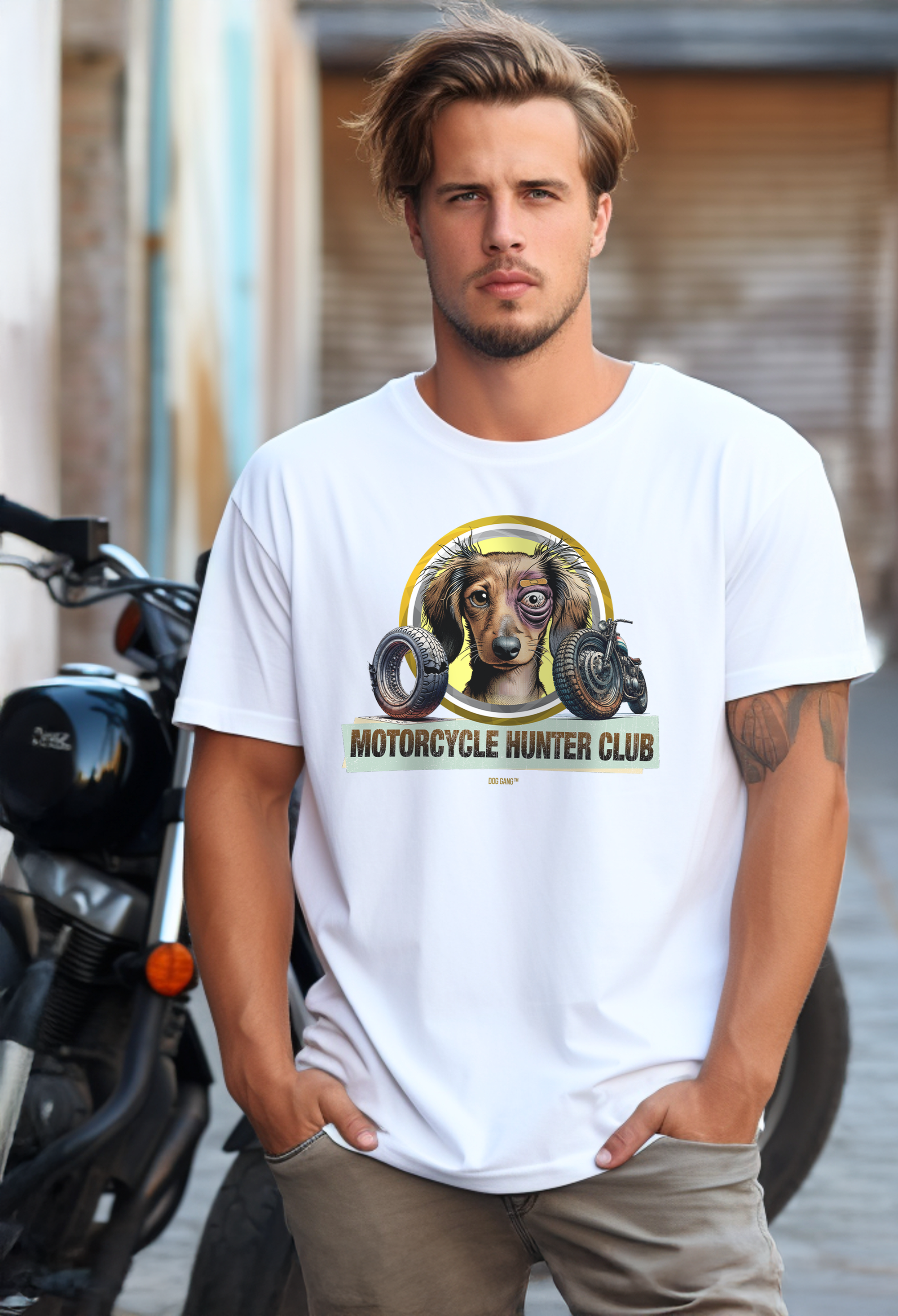 A man with short hair stands on a street in front of a motorcycle. He is wearing a white Dog Gang T-shirt with a Motorcycle Hunter Club motif showing a slightly disheveled but happy dog ​​face with a black eye and bitten motorcycle parts.
