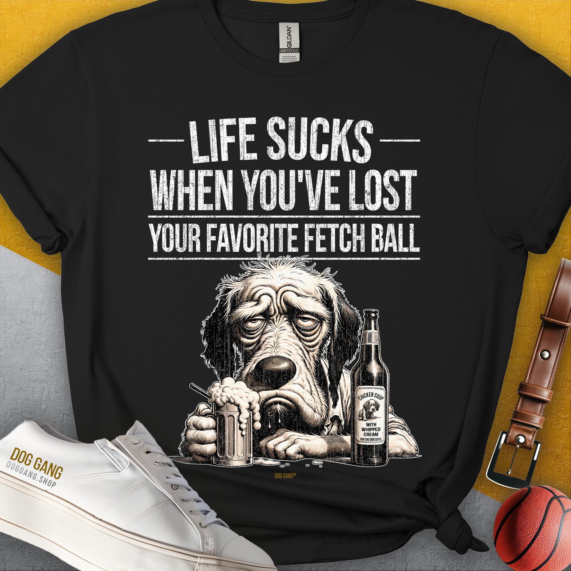 Unisex-T-Shirt mit einem lustig gezeichneten traurigen Hund mit eine Flasche (Flaschentikett: "Chicken Soup with whipped cream, for sad dog days") und dem Text „Life Sucks When You've Lost Your Favorite Fetch Ball“. Design von Dog Gang.