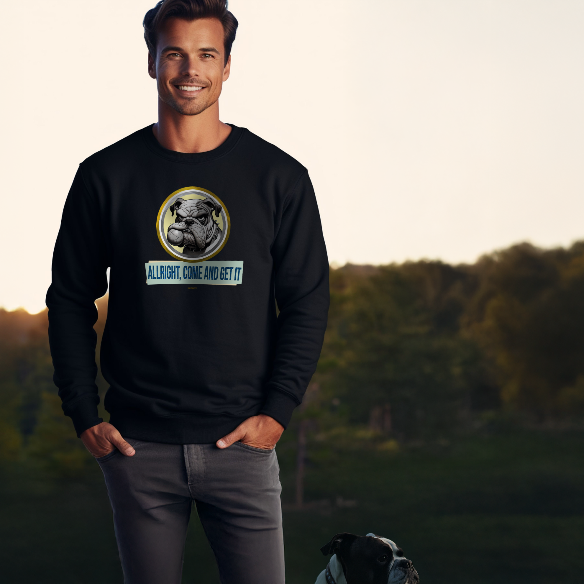 A man stands outdoors, smiling, wearing a black sweatshirt with a cartoon bulldog and the text Alright, Come and Get It. He has his hands in his pockets. A real bulldog is seated nearby on the grass. Trees and a blue sky are in the background.