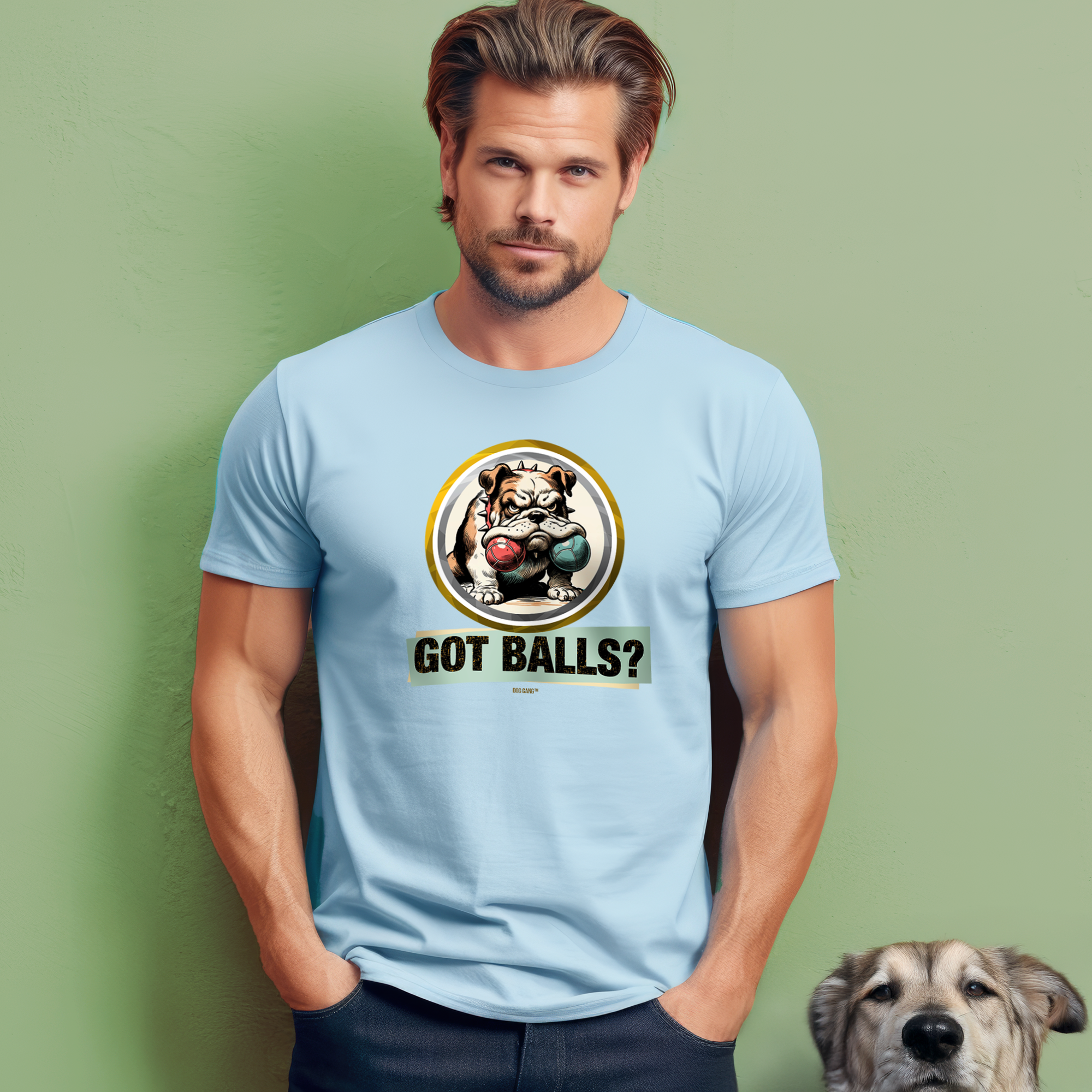 A man with tousled hair stands against a light green wall, wearing a light blue T-shirt featuring a cartoon bulldog with boxing gloves and the text Got Balls? A dog is peeking from the bottom right of the image.