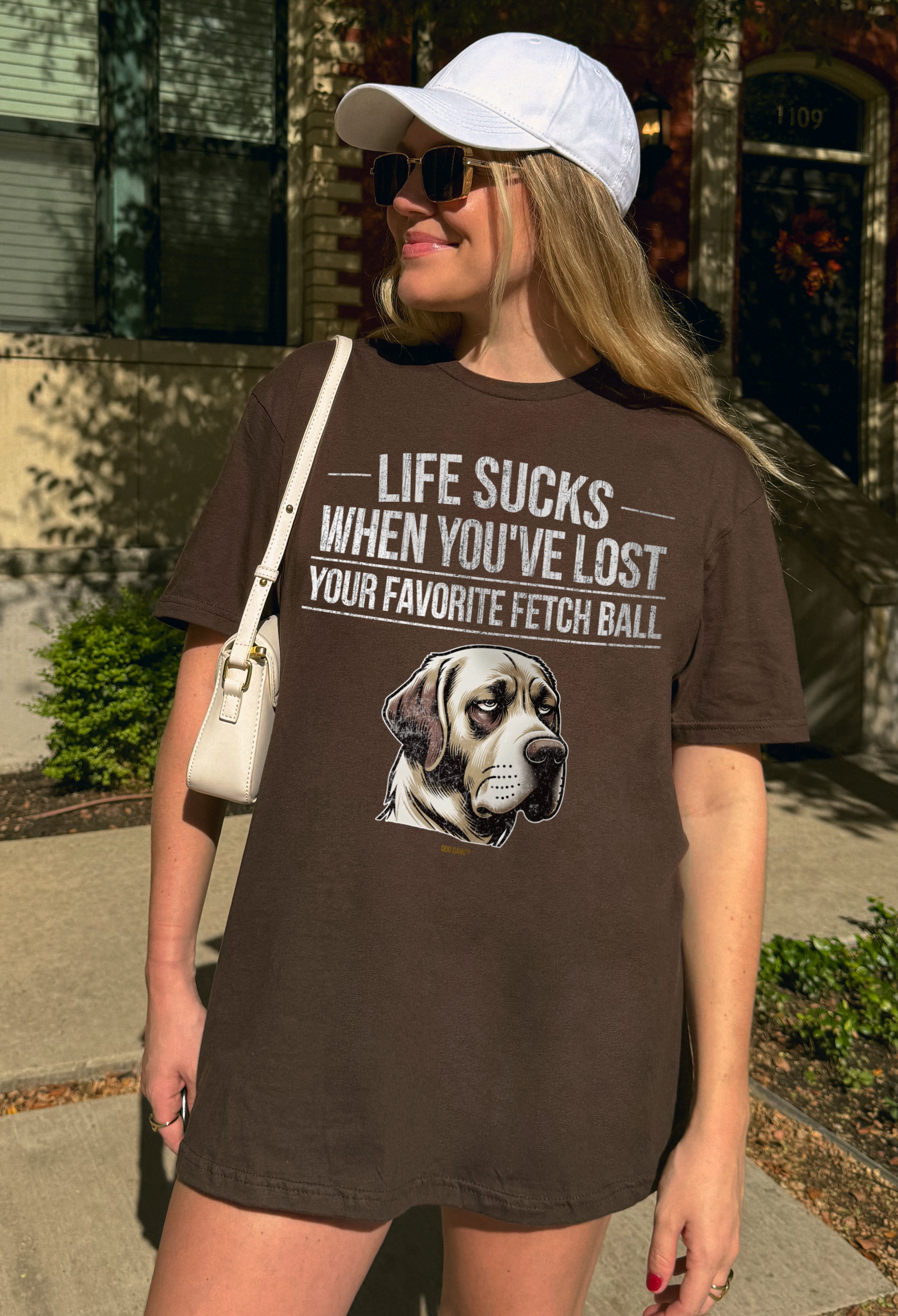 A casual woman with sunglasses and a white cap wears an oversized brown unisex T-shirt by DOG GANG with the text "Life sucks when you've lost your favorite fetch ball" and the funny drawn portrait of a depressed looking Labrador Retriever.